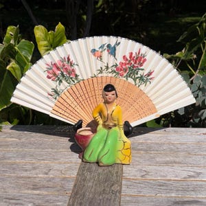 May include: A decorative fan with a wooden handle and a painted design of flowers and birds. A ceramic figurine of a woman in a yellow and green dress is in front of the fan. The fan is open and displayed on a wooden surface.