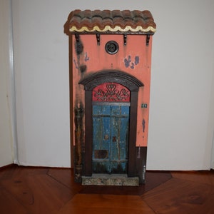 May include: A miniature wooden model of a building with a red tiled roof, a blue door with a metal grate, and a pink facade. The building has a number "87" on the side.