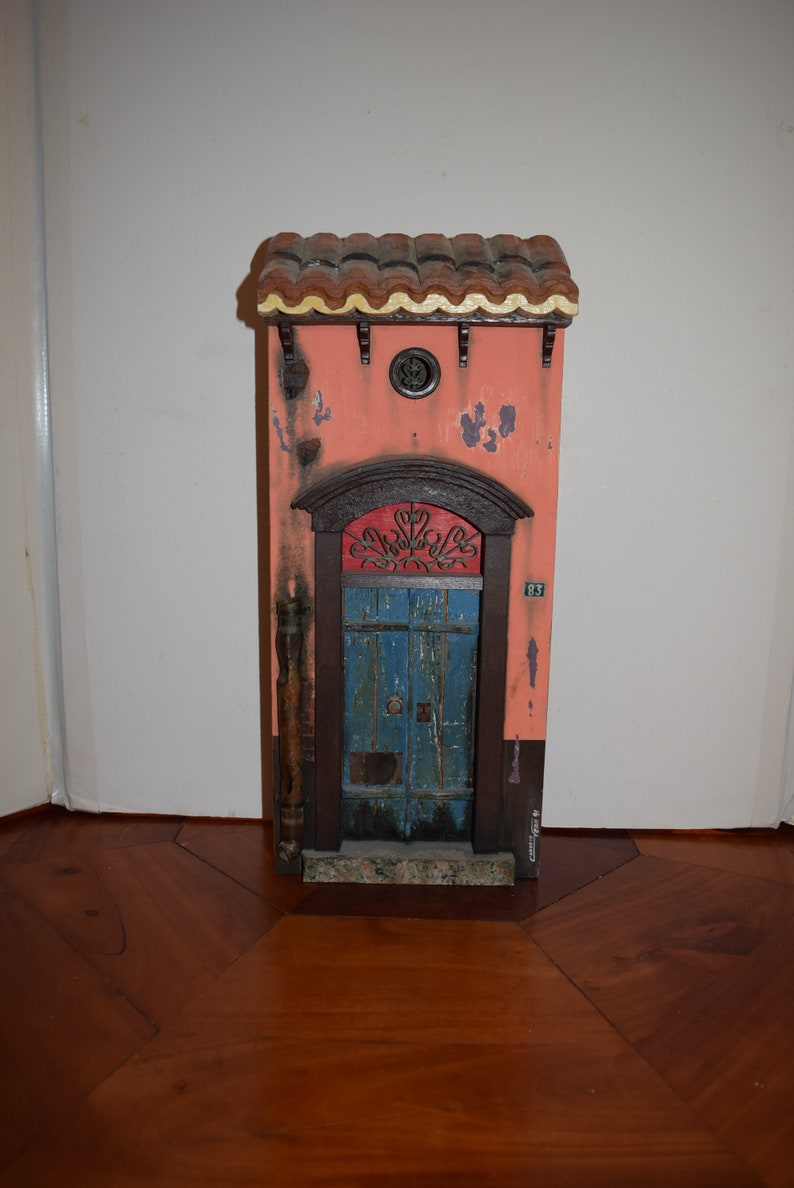 May include: A miniature wooden model of a building with a red tiled roof, a blue door with a metal grate, and a pink facade. The building has a number "87" on the side.