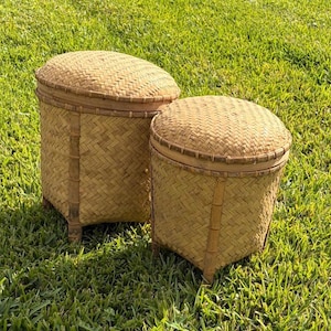 May include: Two woven bamboo storage baskets with lids. The baskets are light brown with a textured weave pattern. They have a cylindrical shape with a rounded top and are set on short legs. The baskets are sitting on green grass.
