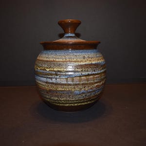 May include: A brown ceramic jar with a lid, featuring a striped pattern in shades of blue, brown, and yellow.