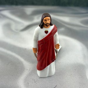 May include: A small figurine of a figure with long brown hair and a beard, wearing a white robe with a deep red sash draped across the chest. A small red heart emblem is visible on the chest area. The figure is attached to a metal ring.