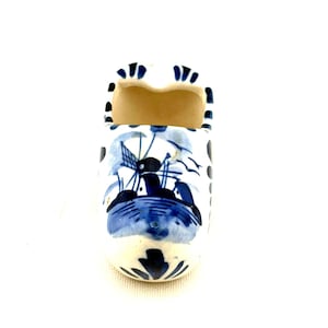 May include: A small, ceramic Dutch clog, painted with a blue and white windmill scene. The miniature shoe has a rounded toe and a hollow interior. Black dots and blue accents decorate the edges. The clog is a decorative item.