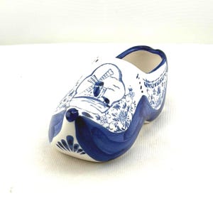 May include: A ceramic Dutch clog, painted white with blue floral and windmill designs. The shoe has a rounded toe and a curved sole, with a dark blue trim along the top edge and base. The clog is a decorative item.