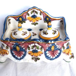 May include: A ceramic soap dish with two matching lidded jars. The dish and jars are white with blue, yellow, and red floral designs. The dish has a scalloped edge and is supported by three small feet.