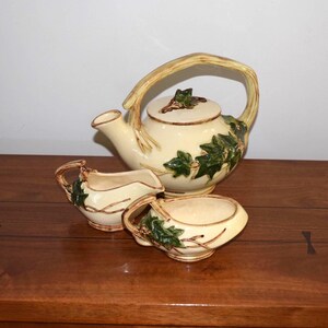 May include: A cream-colored ceramic tea set with a teapot, creamer, and sugar bowl. The set features green ivy leaf detailing and a light brown handle and trim. The teapot has a lid with a leaf-shaped knob.