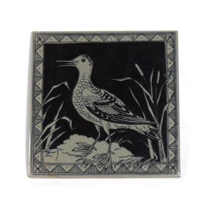 May include: Square ceramic tile featuring a black and white illustration of a bird standing near reeds. The bird has detailed feather patterns and a long beak. The tile has a decorative border with geometric patterns.