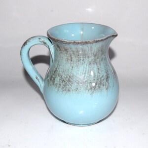 May include: A ceramic pitcher with a glossy light blue glaze and a curved handle. The upper portion of the pitcher features a decorative, brushed dark brown finish that fades into the solid light blue color toward the rounded base.