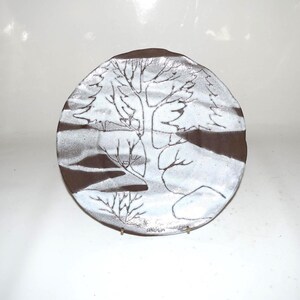 May include: A handcrafted ceramic plate with a winter landscape design. The plate features a brown rim and a white center with a detailed illustration of trees and a small animal. The artwork is signed at the bottom.