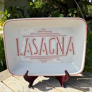 May include: White rectangular baking dish with red trim and the word "LASAGNA" in red, stylized lettering. The dish is supported by a dark red stand, and is set against a backdrop of green foliage.