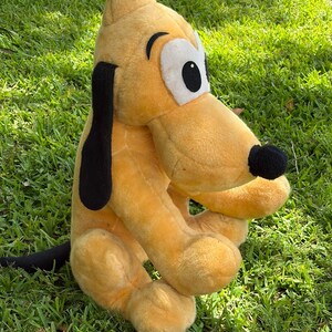 May include: A plush toy of Pluto, a yellow dog with black ears and a black nose, sitting on green grass. The toy has large white eyes with black pupils and is a classic Disney character.