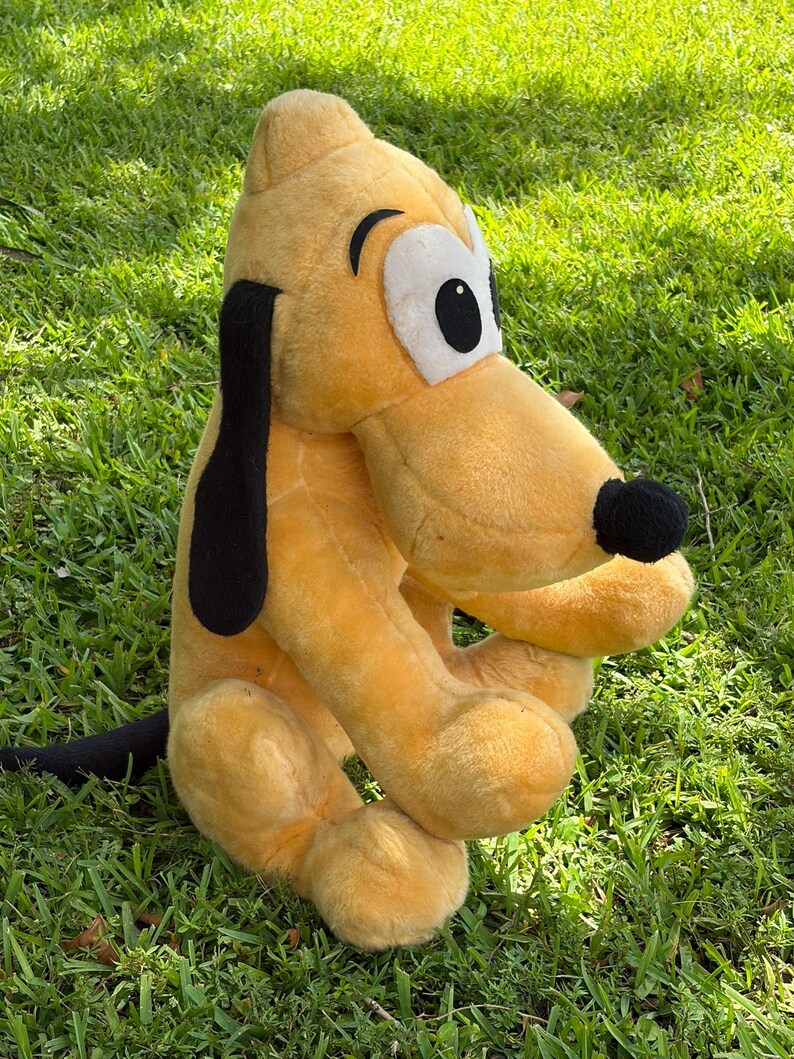 May include: A plush toy of Pluto, a yellow dog with black ears and a black nose, sitting on green grass. The toy has large white eyes with black pupils and is a classic Disney character.