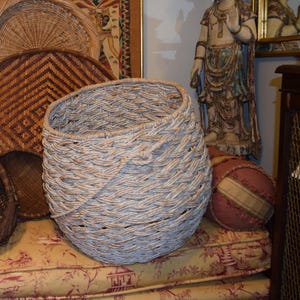 May include: A large, woven basket with a light gray color, set on a floral patterned cushion. The basket has a rounded shape and a textured surface. Other woven items and a statue are in the background.