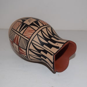 May include: A hand-painted ceramic vase with a geometric design in black, red, and white. The vase is made in a traditional style and has a rounded shape.