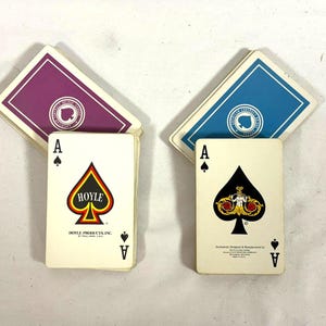 Puede incluir: Dos barajas de cartas con dorsos morados y azules. El As de Picas aparece en el frente de cada baraja. Una baraja tiene el logotipo de Hoyle y la otra un dise&ntilde;o ornamentado. Las cartas est&aacute;n sobre una superficie blanca.