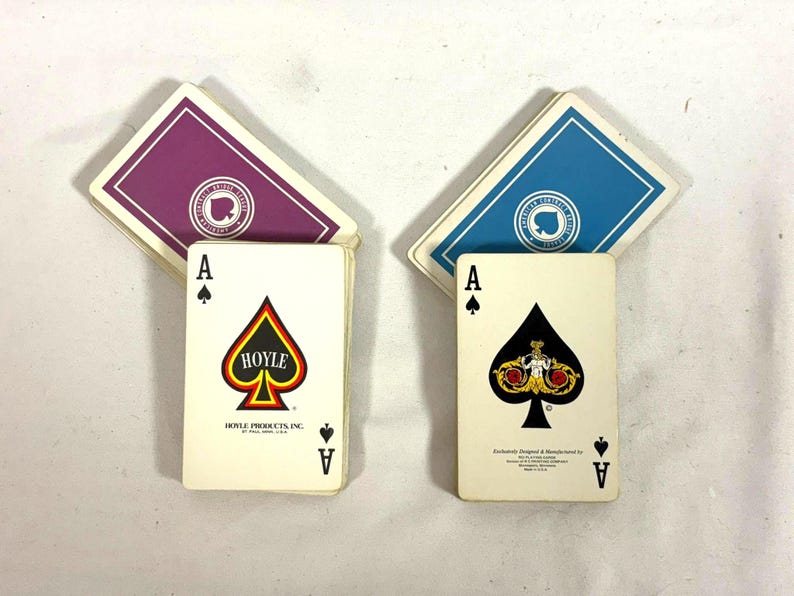 Puede incluir: Dos barajas de cartas con dorsos morados y azules. El As de Picas aparece en el frente de cada baraja. Una baraja tiene el logotipo de Hoyle y la otra un dise&ntilde;o ornamentado. Las cartas est&aacute;n sobre una superficie blanca.