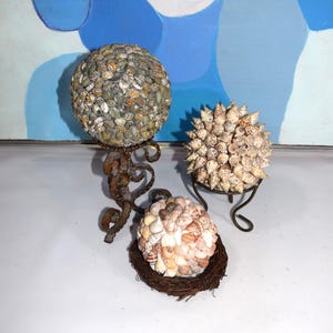 May include: Three decorative spheres made of seashells. One sphere is mounted on a rusty metal stand, another on a black metal stand, and the third rests on a woven brown base. The spheres are various sizes and colors.