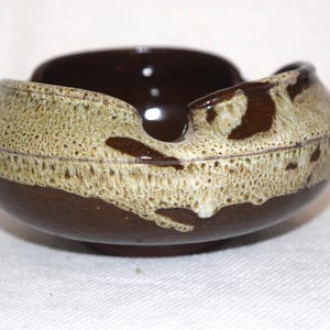 May include: A ceramic ashtray with a dark brown interior and exterior. The rim features a speckled tan and brown glaze. The ashtray has a unique, curved design with a small notch for resting items.