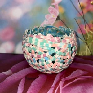 May include: A woven ceramic bowl with a pastel color scheme of pink, blue, and green. The bowl has a round shape and is made of interwoven strips of ceramic. The background features pink flowers and a blurred blue and pink backdrop.