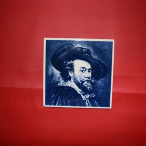 May include: A square ceramic tile featuring a portrait of a man in a large hat, rendered in shades of blue and white. The man has a beard and mustache. The tile is set against a red background.