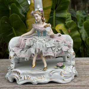 May include: A porcelain figurine of a woman seated on a white bench. The woman wears a blue and pink dress and holds a fan. The base is white with gold accents and a small rose.