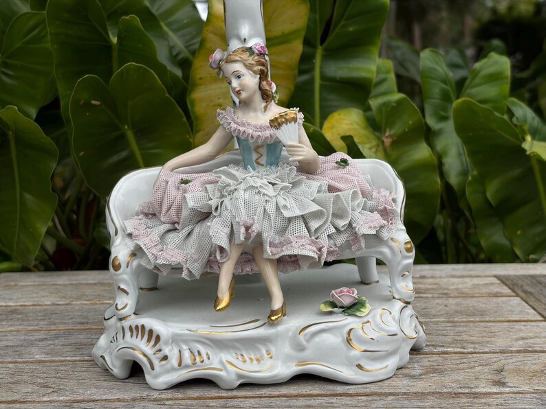 May include: A porcelain figurine of a woman seated on a white bench. The woman wears a blue and pink dress and holds a fan. The base is white with gold accents and a small rose.