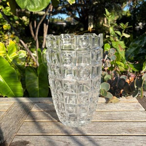 May include: A clear glass vase with a geometric, square pattern. The vase is cylindrical with a slightly flared top. The design features a series of squares and circles, creating a textured appearance. The vase is sitting on a wooden surface.