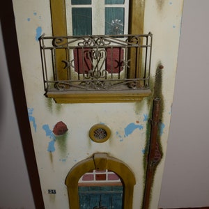 May include: A miniature model of a building with a weathered white facade, a red brick accent, and a blue door with a wrought iron gate. The building has a balcony with a wrought iron railing and a window with a broken pane of glass. The number "21" is painted on the side of the building.