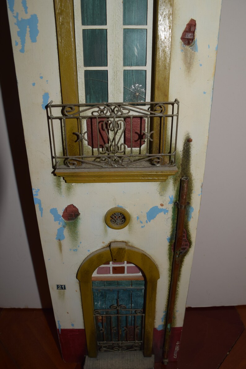May include: A miniature model of a building with a weathered white facade, a red brick accent, and a blue door with a wrought iron gate. The building has a balcony with a wrought iron railing and a window with a broken pane of glass. The number "21" is painted on the side of the building.