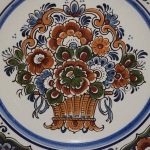 K&ouml;nnte beinhalten: Dekorative Keramikplatte mit einem Blumengestaltungs-Design. Die Platte hat einen wei&szlig;en Hintergrund mit einem blauen Rand und ein zentrales Bild eines Korbes mit orangefarbenen, blauen und gr&uuml;nen Blumen.