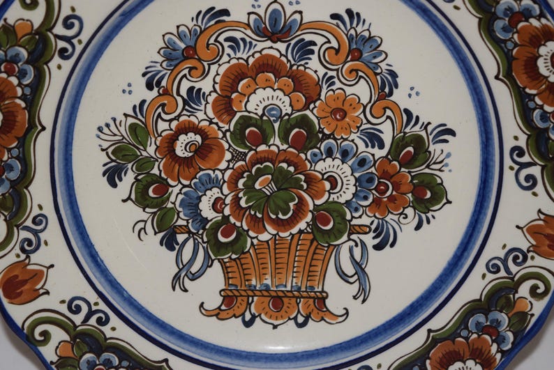 K&ouml;nnte beinhalten: Dekorative Keramikplatte mit einem Blumengestaltungs-Design. Die Platte hat einen wei&szlig;en Hintergrund mit einem blauen Rand und ein zentrales Bild eines Korbes mit orangefarbenen, blauen und gr&uuml;nen Blumen.