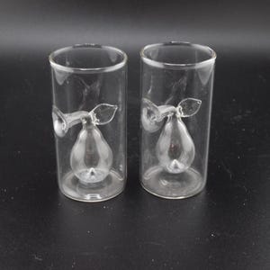 May include: Two clear glass shot glasses, each featuring a pear-shaped design inside. The pears have stems and leaves. The glasses are cylindrical with a flat base, perfect for serving a small drink.