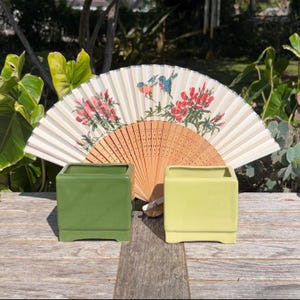 May include: Two square ceramic planters, one in a vibrant green and the other in a soft yellow, sit in front of a decorative fan. The fan features a floral and bird design in shades of red, blue, and beige, adding a touch of elegance.