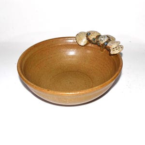 May include: A handmade, brown ceramic bowl with a decorative, textured design on the rim. The bowl has a smooth interior and a slightly rough exterior. The rim features a cluster of small, abstract shapes with blue accents.