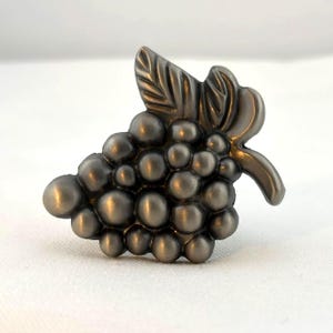 May include: A decorative cabinet knob in the shape of a bunch of grapes. The knob is made of a dark, metallic material with a detailed leaf and stem. The grapes are round and clustered, adding a touch of nature-inspired elegance to furniture.