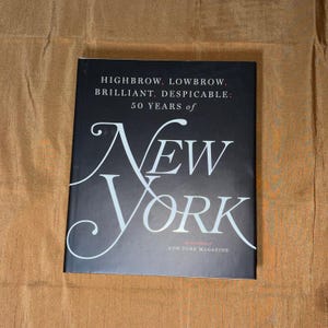May include: A hardcover book with a black cover featuring the title "NEW YORK" in large, elegant white script. The text "HIGHBROW, LOWBROW, BRILLIANT, DESPICABLE: 50 YEARS of" is above the title. The book is on a tan surface.