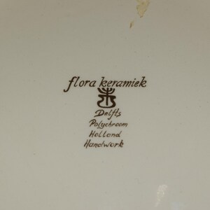 K&ouml;nnte beinhalten: Cremefarbene Keramikoberfl&auml;che mit braunem Text und Logo. Der Text lautet "flora keramiek", "Delfts Polychroom Holland Handwork". Das Logo ist ein stilisiertes Blumendesign.