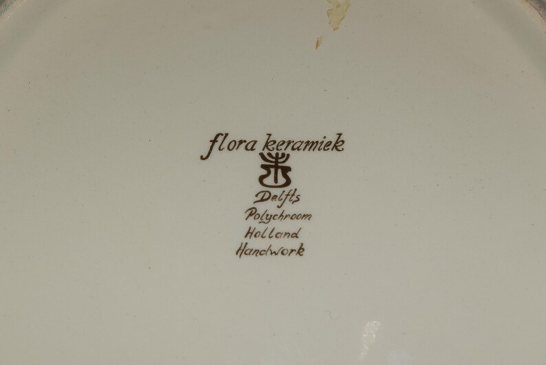 K&ouml;nnte beinhalten: Cremefarbene Keramikoberfl&auml;che mit braunem Text und Logo. Der Text lautet "flora keramiek", "Delfts Polychroom Holland Handwork". Das Logo ist ein stilisiertes Blumendesign.
