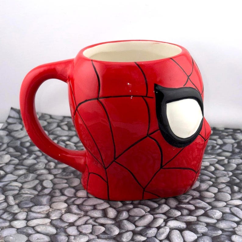 May include: A red ceramic mug shaped like Spider-Man's head, featuring black web lines and a white eye. The mug has a curved handle and a white interior. The mug is sitting on a gray stone surface.