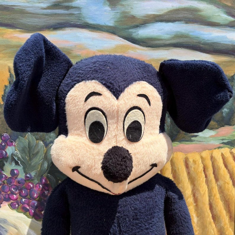 May include: A plush toy of a cartoon mouse with large, dark blue ears and a matching body. The mouse has a cream-colored face, large black eyes, and a black nose. The background features a landscape painting with rolling hills and a vineyard.