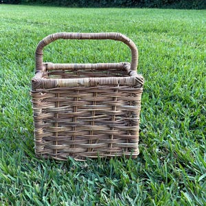 May include: A square, woven wicker basket with a handle. The basket is made of light brown and tan wicker and sits on a bed of green grass. The handle is arched and attached to the top of the basket.