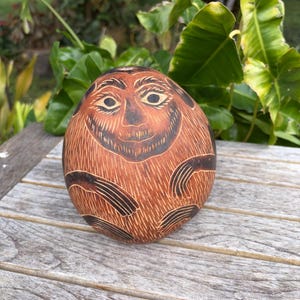 May include: A round, brown gourd featuring an etched, dark-toned illustration of a primate-like face with detailed facial features and limbs wrapped around the body. The surface displays fine, linear carvings that create texture and depth.