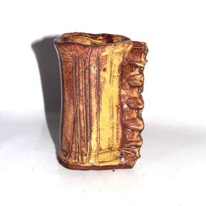 May include: A handmade ceramic vase with a textured surface. The vase features a combination of brown and yellow colors. Vertical lines and a textured edge add to the unique design. The vase is a decorative item.