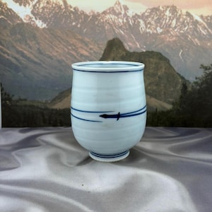 May include: A white ceramic cup with a smooth, rounded body and a slightly flared rim. The exterior features horizontal blue lines and a small, stylized blue arrow graphic. The base is circular and slightly raised, with thin blue line detailing.