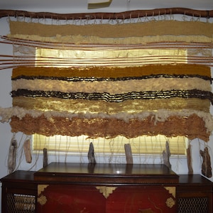 May include: A woven wall hanging with a natural wood frame and a variety of textures and colors, including brown, beige, and white. The hanging is made of different materials, including yarn, fabric, and wood.