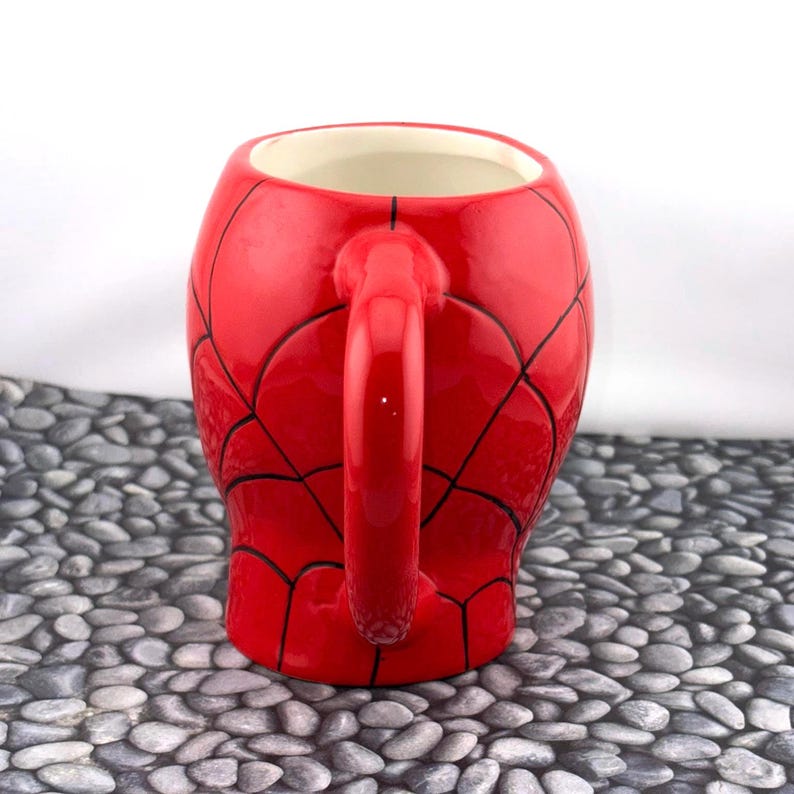 May include: A red ceramic mug with a black spiderweb design, featuring a curved handle. The interior of the mug is white. The mug is a novelty item, likely inspired by the Spider-Man character.