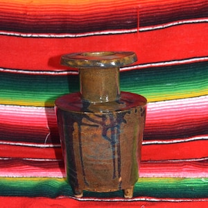 May include: A handcrafted ceramic vase with a dark brown glaze and a cylindrical body. The vase has a flared top and a small, round base with three tiny feet. The glaze has dark blue streaks, and the vase sits on a colorful striped textile.