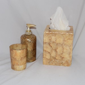 May include: A bathroom set featuring a soap dispenser, tumbler, and tissue box. The set has a warm, neutral color palette with a textured, shell-like appearance. The dispenser has a gold pump, and the tissue box holds white tissues.