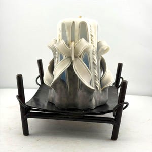May include: A carved pillar candle with a white bow design, set on a black metal candle holder. The candle has a gray base and white accents. The candle holder has a square base and four legs.