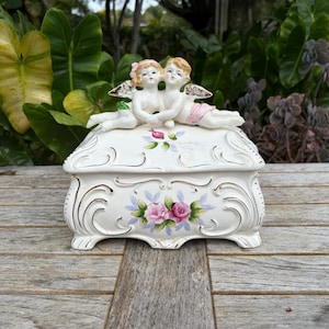 May include: A white ceramic jewelry box with a lid, adorned with pink and purple roses, gold accents, and two cherub figurines on top. The box is set on a wooden surface.